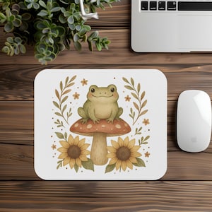 May include: White mousepad featuring a whimsical illustration of a smiling green frog sitting on a red-capped mushroom. The design includes sunflowers, leaves, and small flowers in shades of brown and yellow. A computer mouse and laptop are visible.