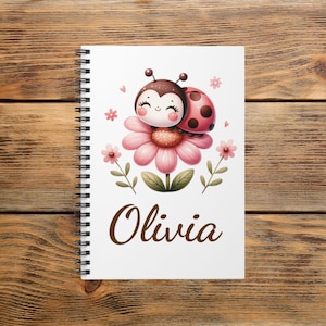 May include: A spiral-bound notebook with a white cover featuring a cartoon ladybug on a pink flower. The ladybug has a smiling face and is surrounded by pink flowers and green leaves. The name "Olivia" is written in brown script below the illustration.