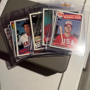May include: A collection of baseball trading cards in protective sleeves. The cards feature players in their team uniforms. One card displays "1984 UNITED STATES BASEBALL TEAM" with "MARK MCGWIRE" and the USA logo. Other cards show "RED" and "PETE".