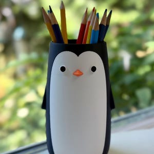Può includere: Un portamatite cilindrico a forma di pinguino con esterno nero, pannello frontale bianco, due piccoli occhi neri, un piccolo becco arancione e due piedini arancioni alla base. Il portamatite è riempito con diverse matite colorate.