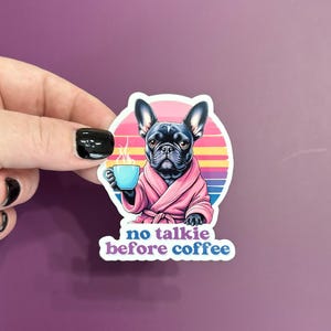 May include: A black and white French bulldog wearing a pink robe and holding a blue mug with steam coming out of it. The background is a colorful sunset with the text "no talkie before coffee".