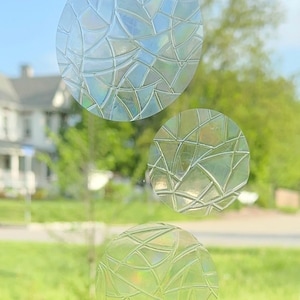 May include: Three round, clear, decorative window clings with a geometric pattern. The clings are applied to a window with a blurred background of green grass and a house.