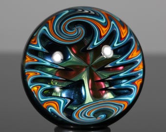 May include: A colorful glass marble with a swirling design in shades of blue, orange, and green. The marble has a glossy finish and two white spots. The design is symmetrical and appears to be a unique piece of art.