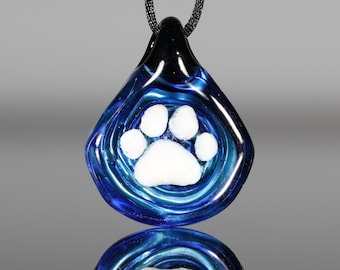 May include: A blue and black glass pendant with a white paw print design. The pendant is teardrop-shaped and hangs from a black cord. The glass has a swirling pattern, and the paw print is centered.