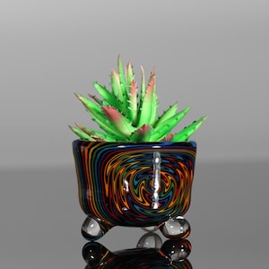 May include: A small, three-legged glass planter featuring a vibrant, multi-colored swirl pattern of orange, blue, and black. The planter holds a faux succulent plant with pointed, light green leaves that have reddish-pink tips at the ends.