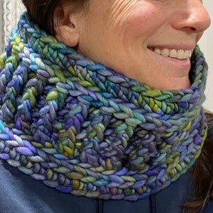 May include: A close-up of a knitted neck warmer in shades of blue, green, and purple. The neck warmer is made of thick yarn and has a chunky texture.