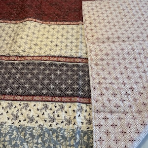 Op de afbeelding: Een doorgestikte sprei met horizontale banen in diverse patronen in de kleuren bordeaux, crème en leigrijs. De stof toont gedetailleerde bloem-, paisley- en geometrische motieven, met een omgeslagen hoek die een witte achterkant met ruitpatroon onthult.