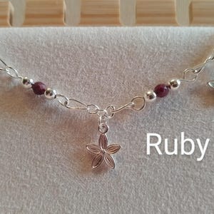 May include: A silver necklace with three flower charms and ruby beads. The necklace has a chain design with a central flower pendant. The word "Ruby" is written in white text.