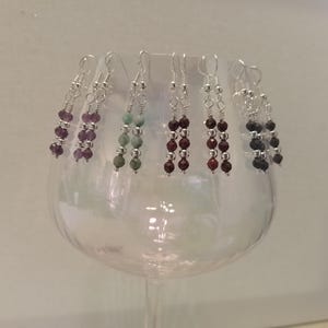 May include: A selection of beaded earrings in shades of purple, green, red, and black. Each earring has a silver hook and small silver beads between the larger, coloured beads. The earrings are displayed on a clear glass.