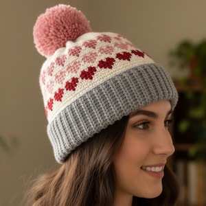 Könnte beinhalten: Handgestrickte Wintermütze mit grauem Rippenbund, weißem Oberteil mit rosa und roten Herzen und einem großen rosa Bommel. Die Mütze ist für Wärme und Stil konzipiert.