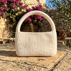 May include: Cream-colored, rectangular crochet handbag with a rounded handle. The bag is made of a thick, textured yarn and has a simple, minimalist design. The bag is sitting on a stone surface, with a background of flowers and a stone wall.