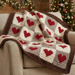 May include: A crocheted blanket with a repeating pattern of red hearts on a cream, grey, and brown background. The blanket is draped over a beige armchair, highlighting its textured design and warm colour palette. The blanket's dimensions are not visible.