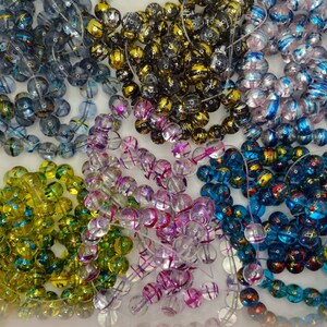 May include: Assortment of colorful glass beads with iridescent finishes. The beads are round and have a variety of colors, including blue, green, pink, and gold.