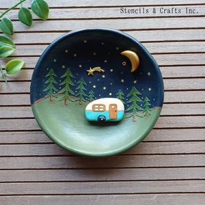 May include: A blue and white camper van with a gold star and crescent moon on a dark blue background with gold glitter stars. The camper van is on a green and brown background with a forest of pine trees. The text "Stencils & Crafts Inc." is in the upper right corner of the image.