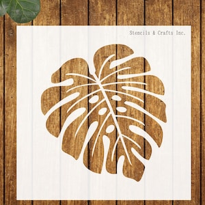 May include: A white stencil featuring a large monstera leaf design. The stencil is on a white background with the text "Stencils & Crafts Inc." in the upper right corner. The background is a wooden surface.