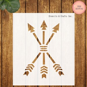 May include: A stencil featuring three arrows pointing in different directions, with a decorative design in the center. The stencil is white and is on a wooden background. Stencils & Crafts Inc.
