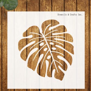 May include: A white stencil featuring a detailed illustration of a Monstera leaf. The stencil is placed on a wooden surface. The text "Stencils & Crafts Inc." is printed in the upper right corner.
