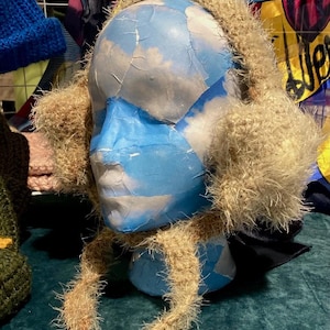 Könnte beinhalten: Ein brauner, flauschiger Ohrenschutz mit einem langen Schal. Der Ohrenschutz befindet sich auf einem Mannequin-Kopf mit einem blau-weiß bemalten Gesicht.