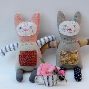 Puede incluir: Dos juguetes de peluche de gato hechos a mano con caras bordadas y detalles de punto. Un gato es rosa y gris con un brazo a rayas blanco y azul, el otro es gris con un brazo a rayas amarillo y rosa. Ambos gatos tienen un bolsillo de punto en el pecho.