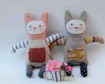 May include: Two handmade plush cat toys with embroidered faces and knitted details. One cat is pink and gray with a white and blue striped arm, the other is gray with a yellow and pink striped arm. Both cats have a knitted pocket on their chest.