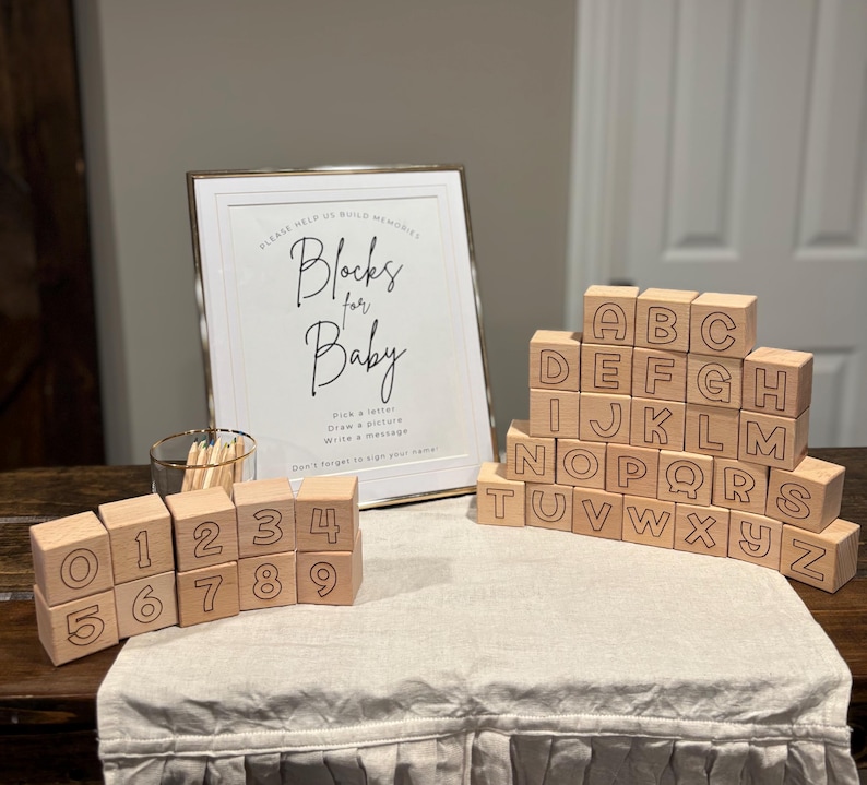 Op de afbeelding: Houten alfabet- en cijferblokken op een tafel. De blokken zijn lichtbruin met gegraveerde letters en cijfers. Op de achtergrond staat een ingelijst bord met de tekst "Blocks for Baby".