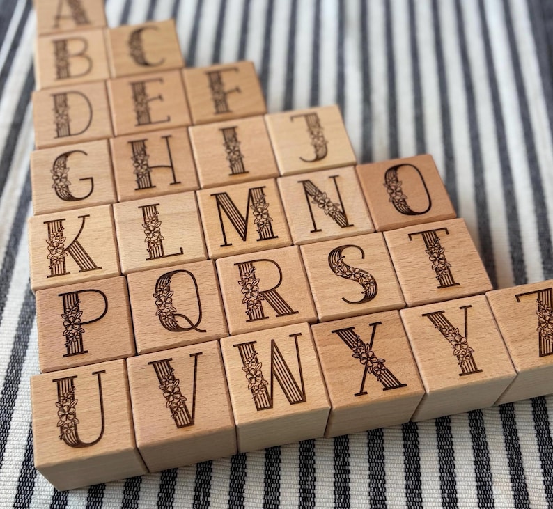 Op de afbeelding: Houten alfabetblokken met gegraveerde letters en bloemaccenten. De blokken zijn in een verspringend patroon gerangschikt en tonen de letters A tot en met Z. De blokken zijn gemaakt van lichtgekleurd hout.