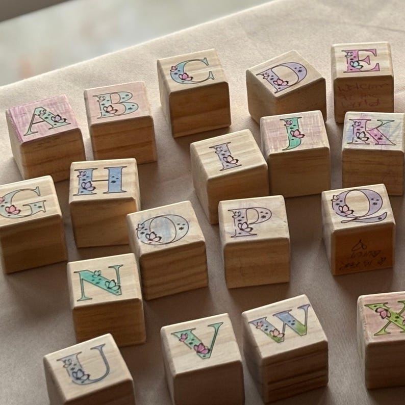May include: Wooden alphabet blocks with pastel floral designs and letters A through X. The blocks are arranged in a random pattern on a light brown surface.