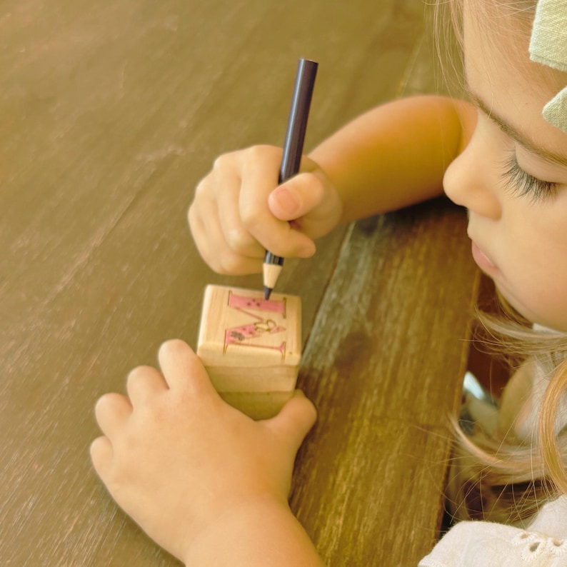 May include: A child's hand is colouring the letter "M" on a wooden block with a pink and white design. The block is sitting on a wooden table. A purple pencil is being used to colour the block.