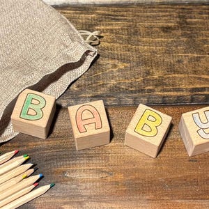 Op de afbeelding: Houten blokken met de letters "BABY" in groene, oranje, gele en grijze kleuren. Een beige trekkoordtas en een set kleurpotloden zijn ook aanwezig op een houten oppervlak. De blokken zijn een klassiek speelgoed.