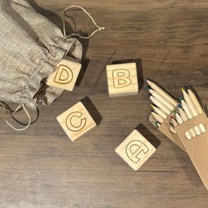 May include: Four wooden blocks with the letters C, D, B, and E engraved on them. The blocks are on a wooden surface with a small hessian bag and a pack of coloured pencils.