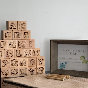Op de afbeelding: Houten alfabetblokken met letters met dinosaurus thema, gestapeld in een piramide. Een ingelijst bordje leest "Versier een blok voor de baby". Potloden liggen voor het frame. De blokken zijn gemaakt van lichtgekleurd hout.