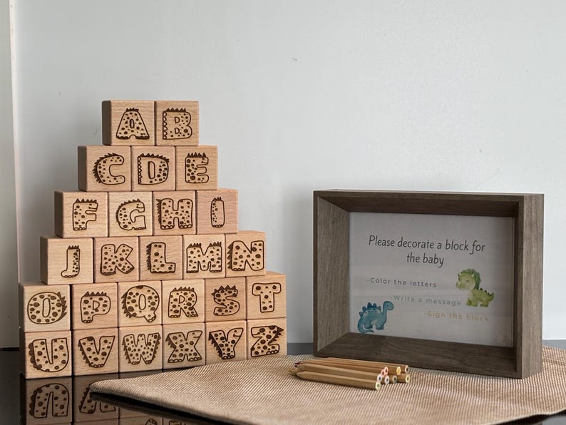 Op de afbeelding: Houten alfabetblokken met letters met dinosaurus thema, gestapeld in een piramide. Een ingelijst bordje leest "Versier een blok voor de baby". Potloden liggen voor het frame. De blokken zijn gemaakt van lichtgekleurd hout.