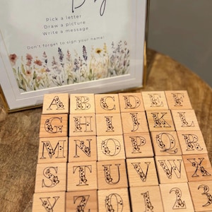 Op de afbeelding: Houten alfabetblokken met bloemmotieven en een ingelijste afbeelding met de tekst: "Kies een letter, Teken een plaatje, Schrijf een bericht, Vergeet niet je naam te ondertekenen!" De blokken liggen op een houten oppervlak.