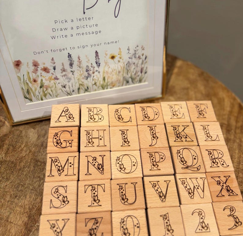 Op de afbeelding: Houten alfabetblokken met bloemmotieven en een ingelijste afbeelding met de tekst: "Kies een letter, Teken een plaatje, Schrijf een bericht, Vergeet niet je naam te ondertekenen!" De blokken liggen op een houten oppervlak.