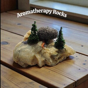 May include: Aromatherapy Rocks. A decorative rock formation with a dark gray porous stone and two miniature green trees. The base is a light-colored, textured rock. The scene includes a small campfire.