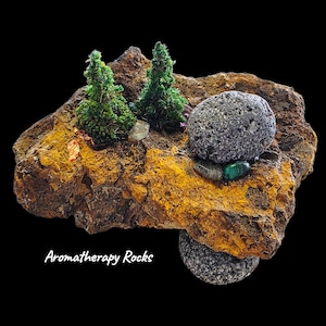 May include: Aromatherapy Rocks display. The display features a textured, multi-colored rock formation with two small green trees and several stones. Two gray, porous stones are stacked on the rock. The text "Aromatherapy Rocks" is written in white cursive.