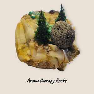 May include: Aromatherapy Rocks. A decorative piece featuring a large, multi-colored rock formation with miniature green trees and a textured, gray stone. The text "Aromatherapy Rocks" is at the bottom.