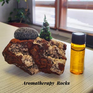 May include: Aromatherapy Rocks display featuring a textured, reddish-brown rock with a small, dark gray stone and a miniature green tree. A small amber glass bottle with a black cap is next to the rock. The text "Aromatherapy Rocks" is at the bottom.