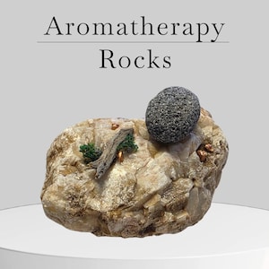 May include: Aromatherapy Rocks are displayed on a white surface. The rocks are beige and tan with a dark gray, textured rock on top. There is a small piece of wood and green foliage attached to the rock. The text "Aromatherapy Rocks" is at the top.