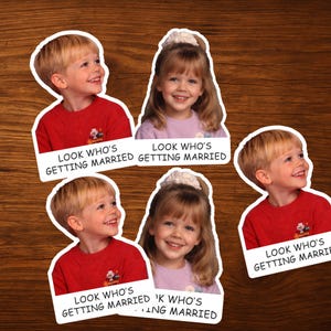 May include: Stickers with images of children and the text "LOOK WHO'S GETTING MARRIED". The stickers depict a boy in a red top and a girl in a lilac top, both smiling. The stickers are on a wooden surface.