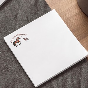 May include: White square notepad with the text "Love is a four legged word" and illustrations of a horse and a dog. The notepad is on a dark gray surface, with a laptop and pen in the background.