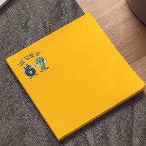 May include: A bright yellow sticky note pad with the text "THE YEAR OF 61" and a cartoon graphic. The pad is square and sits on a dark gray surface, near a laptop and a pen.