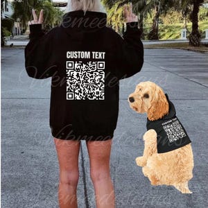 Puede incluir: Una sudadera negra con el texto "CUSTOM TEXT" y un código QR impreso en la espalda. Un perro pequeño que lleva una sudadera negra a juego con un código QR está de pie junto a la persona que lleva la sudadera.