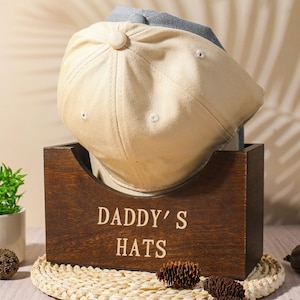 Puede incluir: Una gorra de béisbol beige y azul descansa en una caja de madera. La caja está grabada con las palabras "DADDY'S HATS". Una pequeña planta en maceta y objetos decorativos están en primer plano. El fondo tiene un tono neutro con sombras.