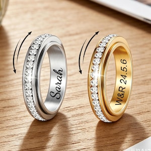 Puede incluir: Dos anillos, uno plateado y otro dorado, cada uno con una banda de piedras transparentes. El anillo plateado tiene grabado el nombre "Sarah". El anillo dorado tiene grabadas las iniciales "W&R 24.5.6". Ambos anillos tienen una banda exterior giratoria.