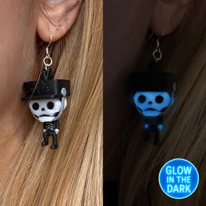May include: A pair of glow-in-the-dark earrings featuring a skeleton wearing a hat. The earrings are silver and the skeleton is black and white. The text "GLOW IN THE DARK" is visible in the bottom right corner of the image.