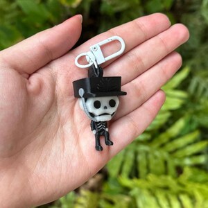 May include: A black and white skeleton keychain with a black hat and a silver clip. The skeleton is wearing a black uniform and has a white skull.