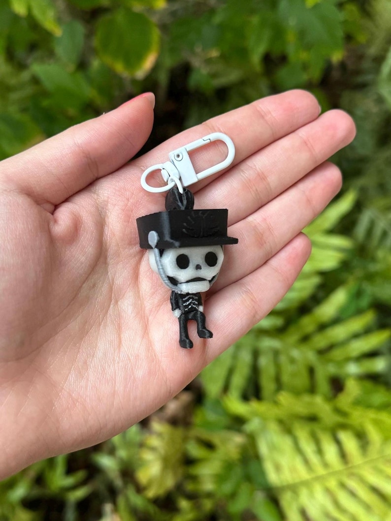 May include: A black and white skeleton keychain with a black hat and a silver clip. The skeleton is wearing a black uniform and has a white skull.