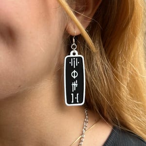 May include: Black rectangle earrings with white symbols, including a circle with a line through it and vertical lines. The earrings are hanging from a person's ear.