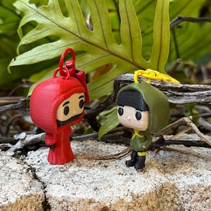 May include: Two miniature figurines, one red and one green, stand side-by-side on a stone surface. The red figure wears a hooded robe, while the green figure is dressed in a military-style outfit. Both have black boots and yellow accents. Keychains are attached.